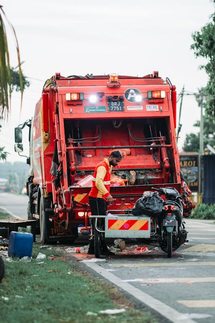 Two large black plastic garbage bags, filled with waste, are situated on a concrete pavement near a low curb in an outdoor setting. One bag appears slightly torn with visible creases and crumples, and