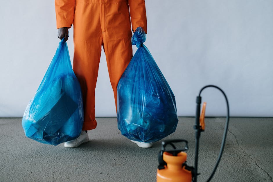 A close-up view of several black plastic rubbish bags filled with waste, placed against a plain, light grey wall. The bags are shiny, slightly crinkled, and tightly tied at the top, with some showing 